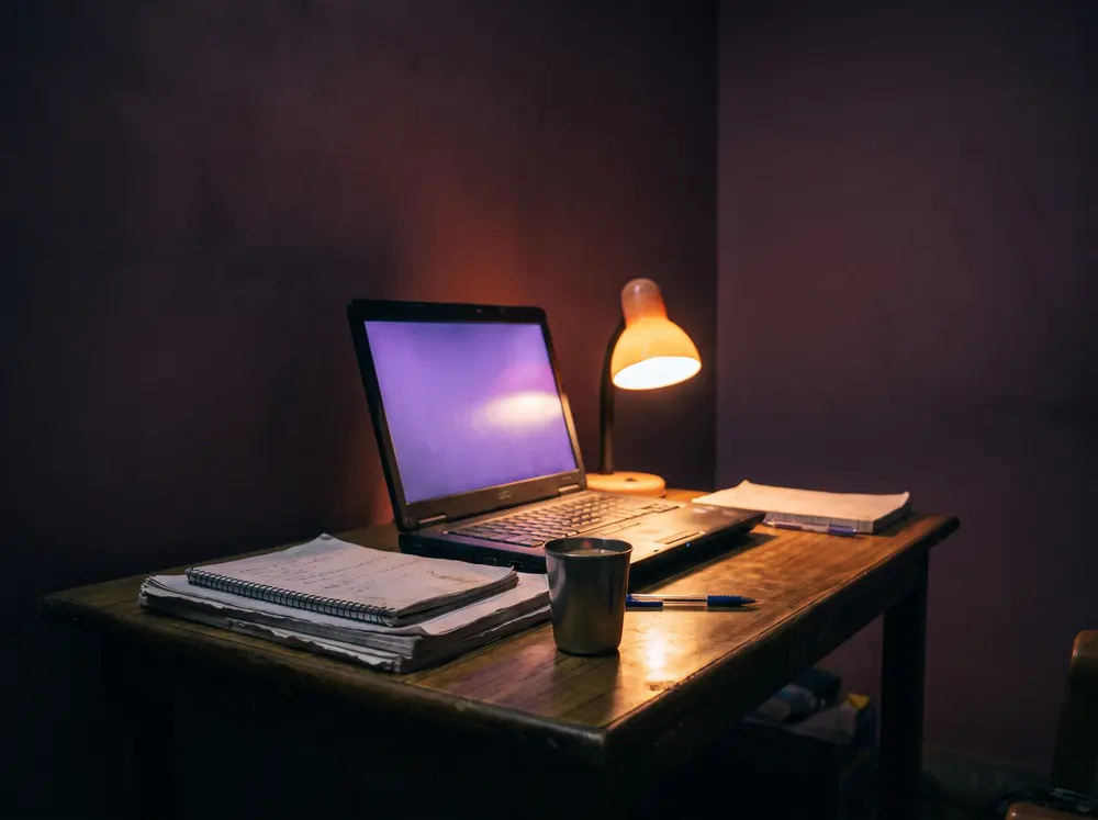 A home study table at night with an older laptop and notebooks.